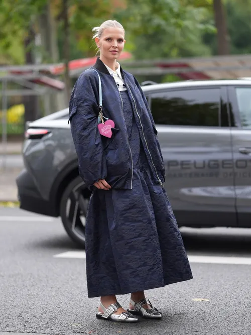 A guest wears silver earrings, white collar dark blue black pattern midi dress, dark blue black pattern oversized jacket, shiny silver metallic Ganni sandals recycled leather shoes, outside Ganni, during the Paris Fashion Week Spring/Summer 2025 on September 24, 2024 in Paris, France.