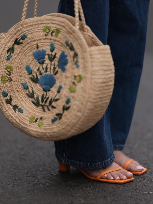 Aline Kaplan trägt eine Jeanshose mit einer bunten Korbtasche und orangefarbenen Sandaletten.
