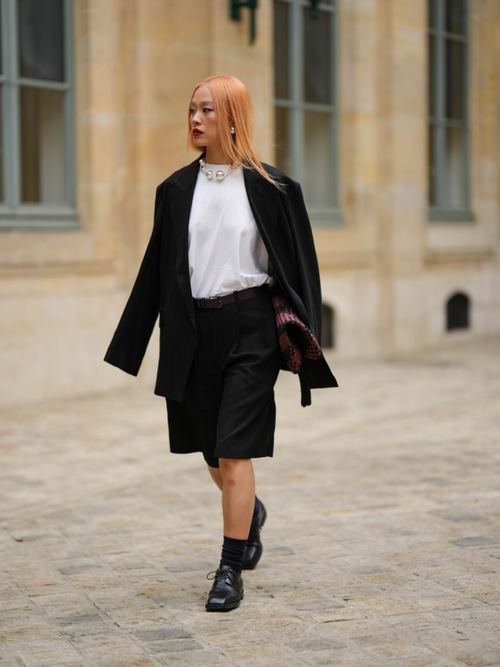 Niki Wu Jie wears a large silver pearls necklace, a white t-shirt, a black blazer jacket, a dark brown shiny leather belt, black suit shorts, a red and black shiny leather braided clutch, black socks, black shiny leather derbies, a gold and silver pearls feet bracelet , outside the COS show, on April 26, 2023 in Paris, France.