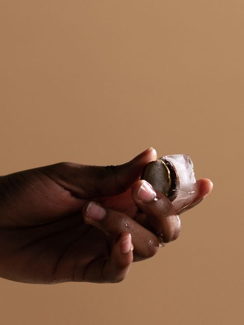 Eine Hand hält einen Eiswürfel in der Hand, vor einem beigen Hintergrund.