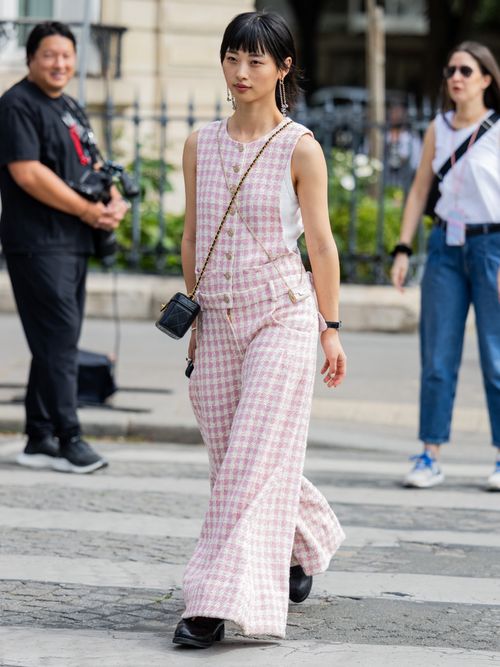A Paris Fashion Week guest wears silver jewellery, a white t-shirt, a pink tweed jumpsuit, a black leather micro bag, and black sneakers.
