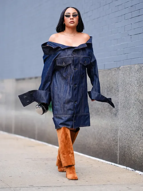 A guest wears silver sunglasses, navy blue off the shoulder oversized buttoned up long sleeve denim jean over the knee dress, bright light brown suede heeled boots, black and silver chrome circle leather bag, outside Area, during the New York Fashion week Spring/Summer 2025 on September 06, 2024 in New York, New York.