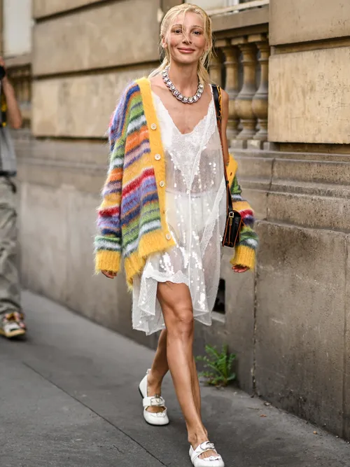 Street Style Look von der Paris Fashion Week, wo ein Gast einen bunten Cardigan, ein transparentes Spitzenkleid, Statementkette und weiße Ledersandalen trägt.