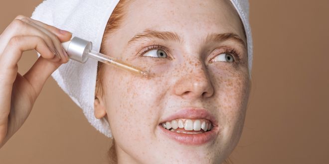 Frau im Badezimmer mit Handtuch um den Kopf gewickelt trägt gerade ein Serum auf die Gesichtshaut auf