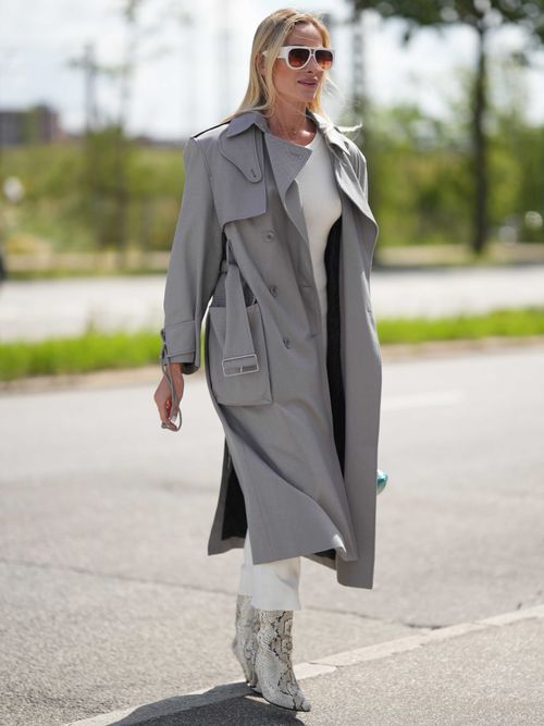 Street Style Look von der Copenhagen Fashion Week, wo ein Gast einen grauen Trenchcoat mit weisem Kleid und Boots in Schlangenprint trägt. Dazu eine weiße Sonnenbrille.