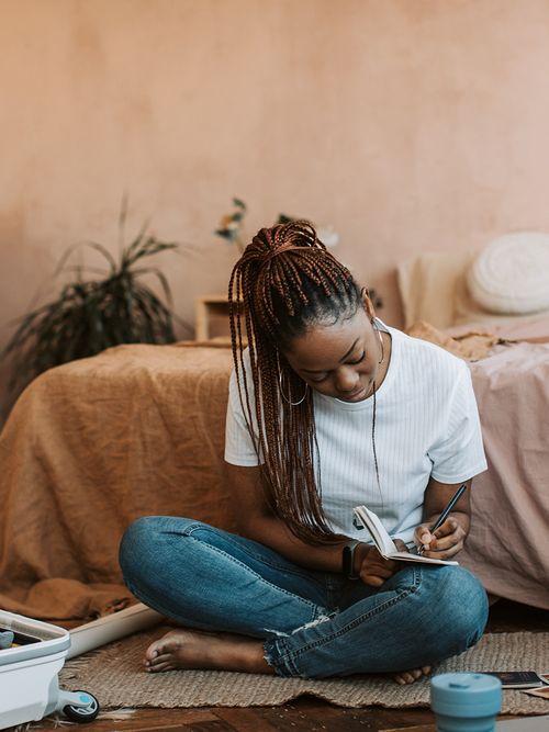 Frau sitzt vor dem Bett und schreibt in ein kleines Notizbuch.