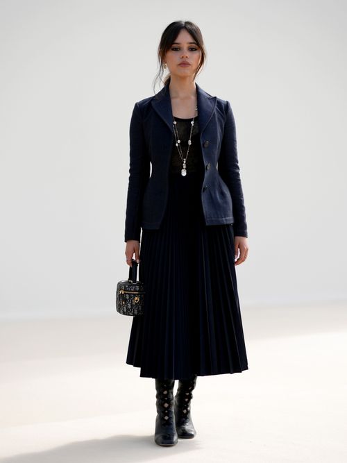 Jenna Ortega wears a necklace, a black top, a navy blue jacket with shoulder pads, a Dior monogram bag, a pleated skirt, black leather boots, outside Dior, during the Womenswear Spring/Summer 2024 as part of Paris Fashion Week on September 26, 2023 in Paris, France.