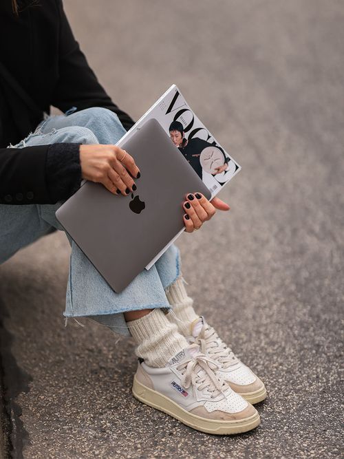 Close up einer Frau mit Jeans, Tennissocken und Sneakers von Autry, die ein MacBook und eine Vogue in der Hand hält.