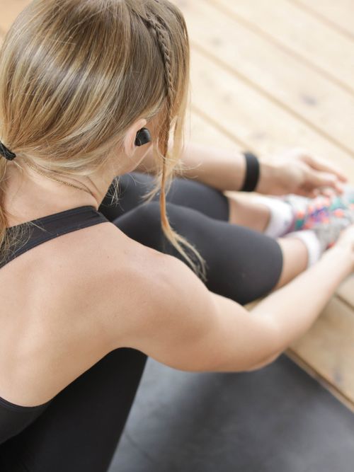 Frau in schwarzen Tanktop und grauer Leggings sitzt auf Yogamatte und bindet sich die Schuhe zu.
