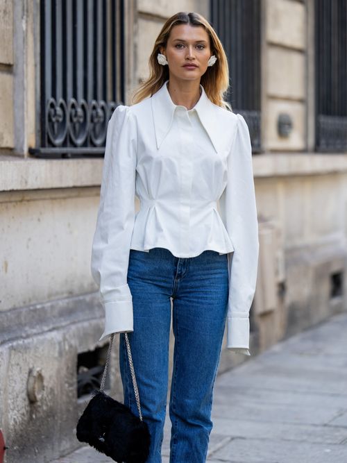 Chloe Lecareux trägt eine weiße Bluse, Statement-Ohrringe, eine Jeans und eine schwarze Chanel Tasche in Paris.