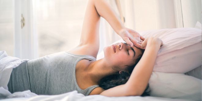 Frau mit grauem Tanktop liegt im Bett und greift sich auf den Kopf.