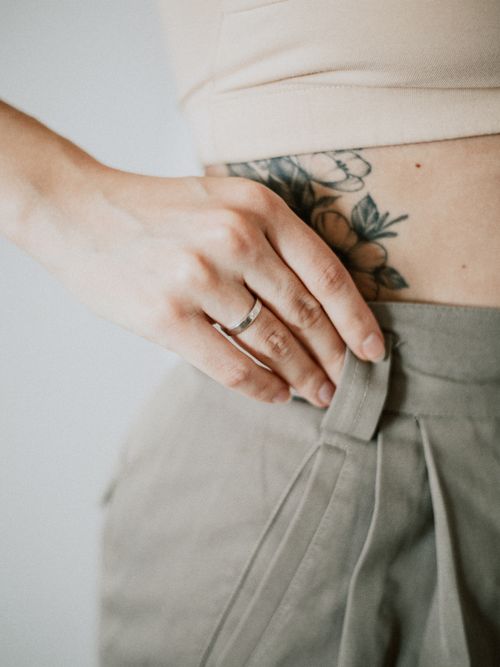Eine Frau zeigt ihr Tattoo am Bauch und trägt eine beige Hose und Sport-Oberteile mit einem Ring am Finger.
