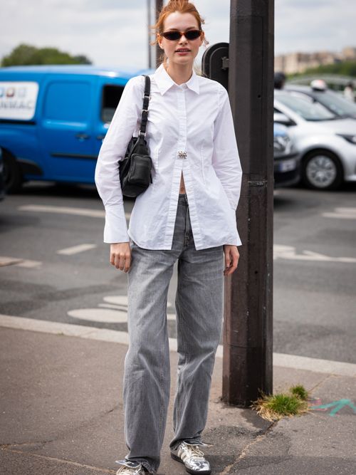 Street Style Look von der Paris Fashion Week, wo ein Model eine weiße Bluse, graue Jeans, schwarze Tasche und silberfarbene Adidas Originals Sneaker trägt.