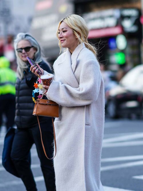 Streetstyle mit einem weißen Mantel, einer braunen Tasche und einem Handy mit Phone Charme in der Hand.