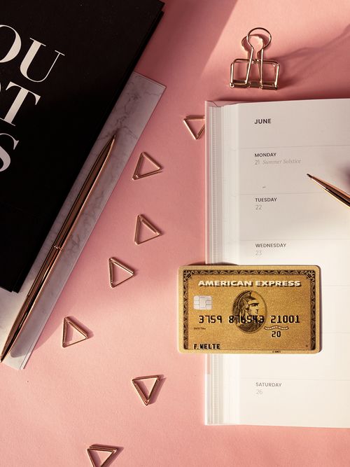 Ein Kalender und goldene Büroklammern liegen gemeinsam mit einer goldenen American Express Karte auf einem pinken Tisch.