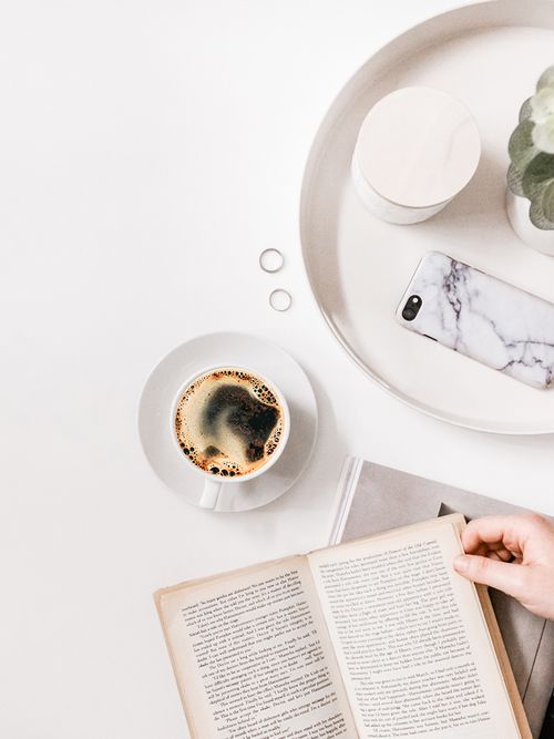 A monochrome white table setting with a round white tray, a plant, a white smartphone, a cup of coffee, and a book.