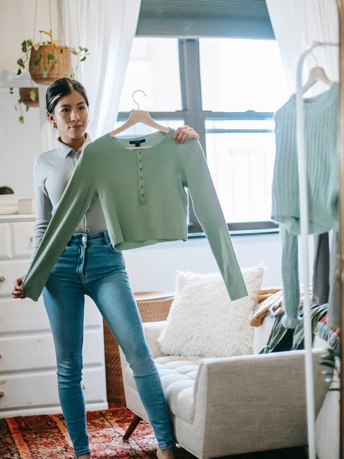 Frau in Jeans hält einen grünen Cardigan in der Hand und betrachtet sich im Spiegel.