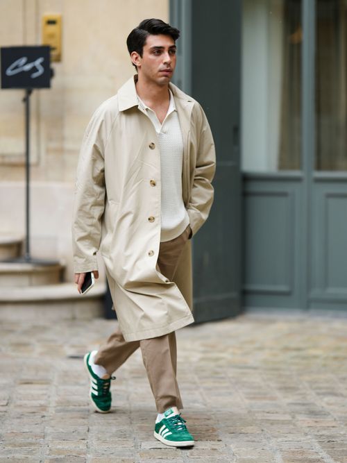 Street Style Look in Paris, wo ein Mann einen beigen Trenchcoat, weißes Strickpolo, beige Hose und Adidas Sneaker trägt.