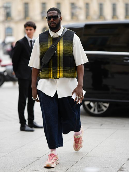 A guest wears black sunglasses, a white ribbed embossed striped print pattern / short sleeves shirt, a black and yellow checkered print pattern wool sleeveless gilet, a black shiny leather crossbody bag from Prada, silver large rings, navy blue denim large short pants, pale pink ribbed socks, pale yellow suede / pink fabric laces sneakers from New Balance , outside the COS show, on April 26, 2023 in Paris, France.