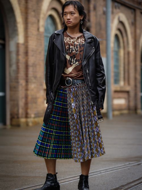 Yuzhen Wang wears a black leather jacket, a brown t-shirt, a midi plaid skirt with leather and metal belt details, and black biker ankle boots.