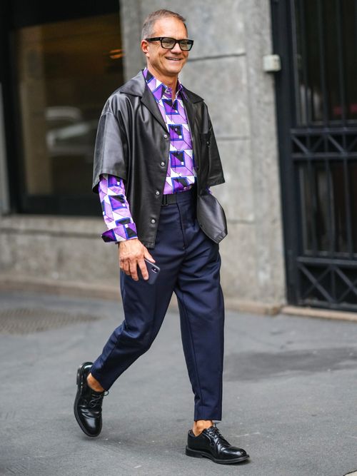 Street Style Look von der Milan Fashion Week, wo ein Gast ein schwarzes Lederhemd mit buntem Shirt darunter trägt. Dazu eine blaue Hose mit schwarzen Schnürschuhe.