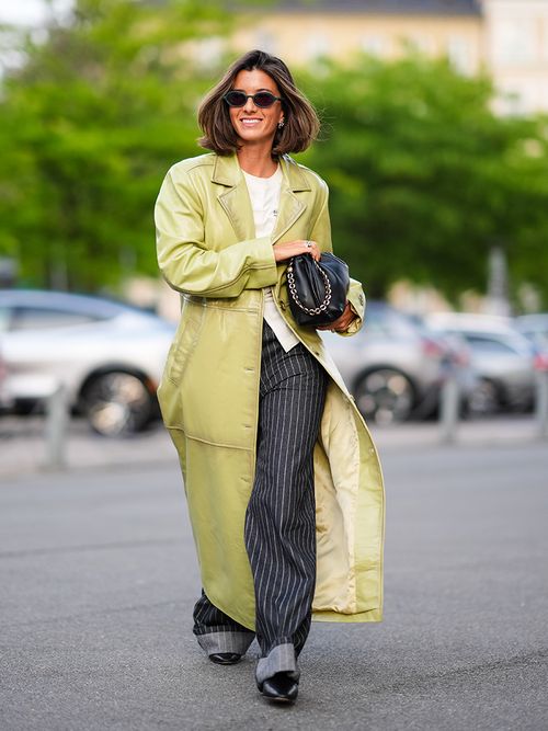 A guest wears black sunglasses, gold earrings, white shirt, shiny light green leather trench coat, black and white lined pants, shiny black leather bag, outside Baum und Pferdgarten, during the Copenhagen Fashion Week Spring/Summer 2024-2025 on August 7, 2024 in Copenhagen, Denmark.