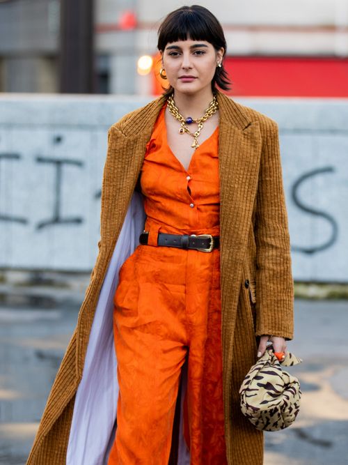 Maria Bernad trägt ein orangefarbenen Overall mit beigen Mantel und Statementkette.
