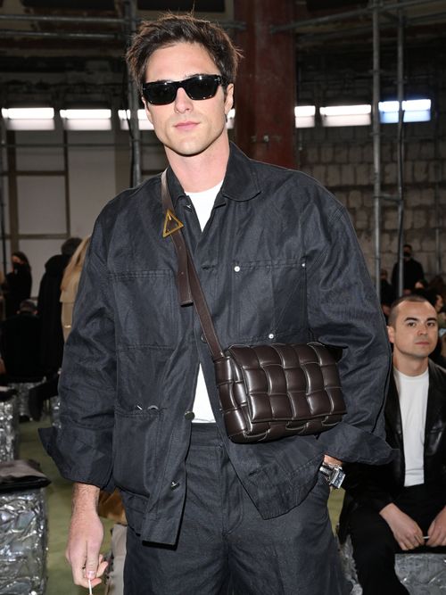 Jacob Elordi attends Bottega Veneta Fashion Show Fall/Winter 2022/2023 wearing square sunglasses, a white t-shirt, a black nylon jacket with matching trousers, and a brown leather bag.