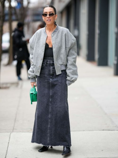 A NYFW guest wears black sunglasses, a pale grey wool teddy coat, a heart-neck pinstripe corset top, a dark wash long denim skirt, a green leather handbag, and pointed black boots.