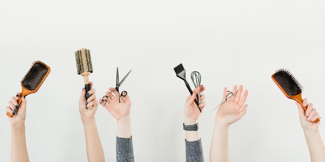 Frauenhände, die eine Bürste, eine Haarschneidescher und einen Färbepinsel in die Luft halten.