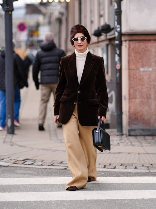 A guest wears a brown fluffy hat, sunglasses, a white turtleneck pullover , a brown oversized velvet blazer jacket , beige flared pants , outside The Garment, during the Copenhagen Fashion Week AW24 on January 31, 2024 in Copenhagen, Denmark.