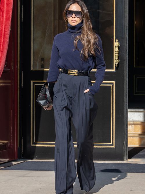 Victoria Beckham trägt einen dunkelblauen Rollkragenpullover mit schwarzer Marlenehose mit schwarzen Gürtel und Golddetails.