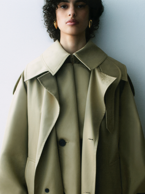 A closeup of a woman with short, curly black hair wearing a beige trench coat.