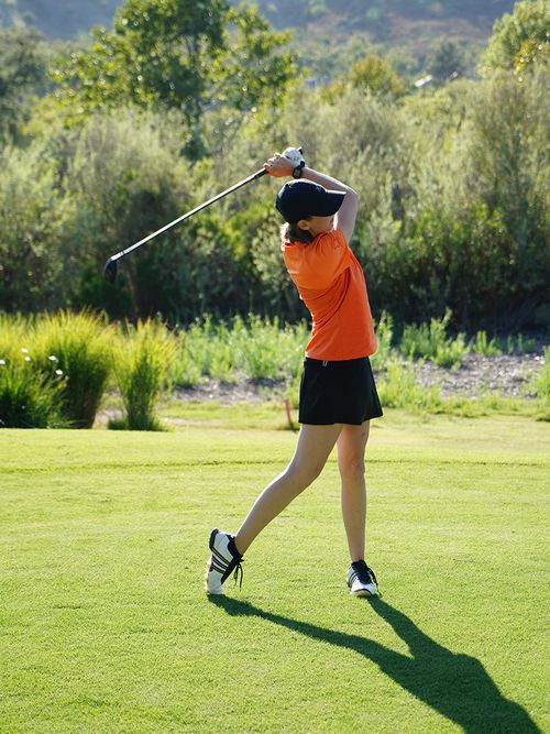 Frau in kurzer Hose, orangenem Shirt am Golfen.
