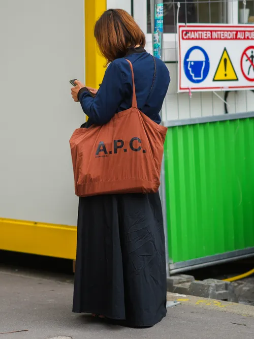 A guest wears a navy blue bomber jacket, a black long skirt, an orange large APC shopping shoulder bag, outside COOL TM, during Paris Fashion Week - Menswear Spring/Summer 2022, on June 23, 2021 in Paris, France.