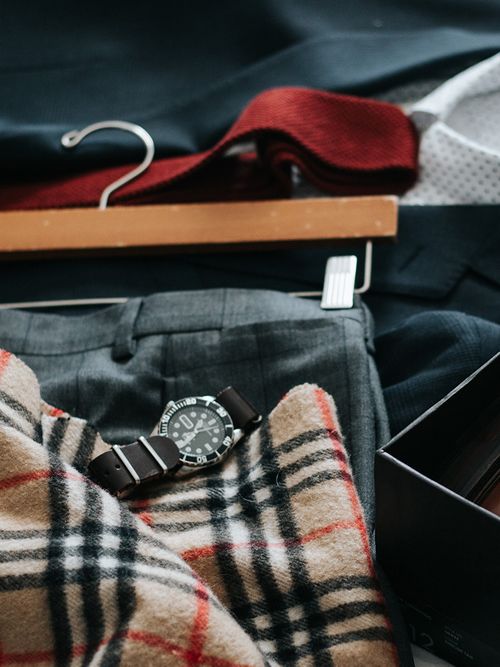 Clothes (a grey suit, a white shirt, a red tie, a Burberry scarf, and a watch) laid flay on a bed.