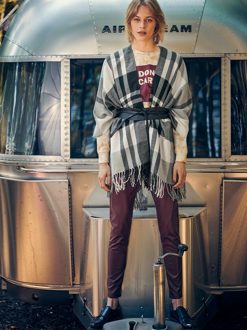 Frau mit roter Lederhose und einem karierten Wollplaid steht vor einem silbernen Wohnwagen.