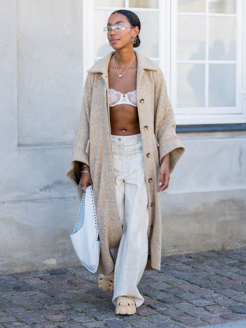 Amaka Hamelijnck trägt einen beigen Mantel, Spitzen-BH, weiße Hose, Sandalen, Sonnenbrille und Tasche in Kopenhagen.
