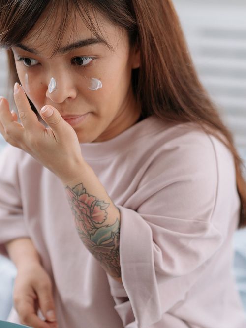 Frau in Rosa T-Shirt und mit einem Tattoo auf der Hand trägt gerade Creme auf ihr Gesicht auf.
