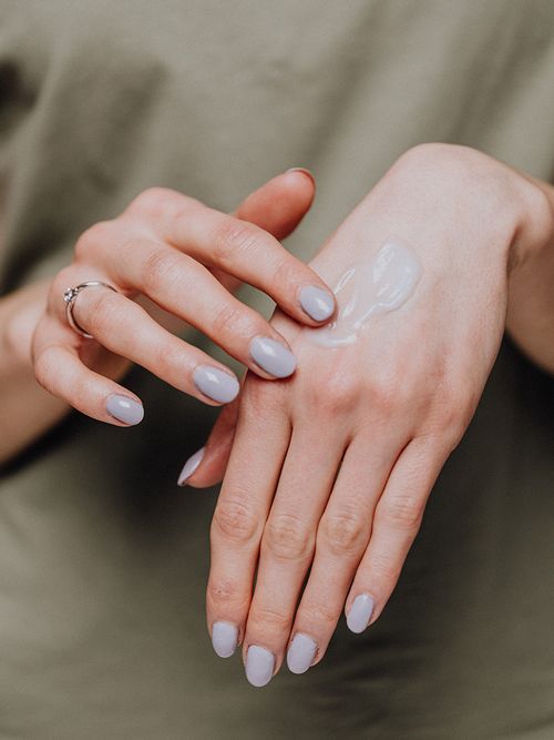 Hände mit einem lila Nagellack die auf den Handrücken eine Creme auftragen.