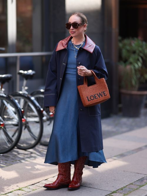 Sonia Lysons seen wearing Loewe brown sunglasses, Pandora gold earrings and necklaces, Baum & Pferdgarten blue long coat with burgundy shiny leather collar, Baum & Pferdgarten blue long dress, Pandora gold and silver rings, Loewe cognac brown raffia logo bag and Ganni red leather cowboy boots, during day three of the Copenhagen Fashion Week (CPHFW) SS25 on August 07, 2024 in Copenhagen, Denmark.