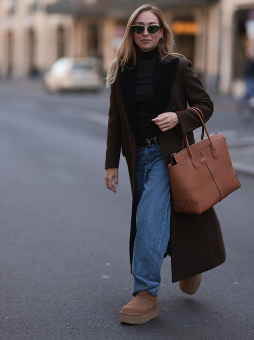 Sonia Lyson trägt einen braunen Mantel mit schwarzem Rollkragen, breiter Jeanshose mit Ugg Boots und einer Tods Tote Bag
