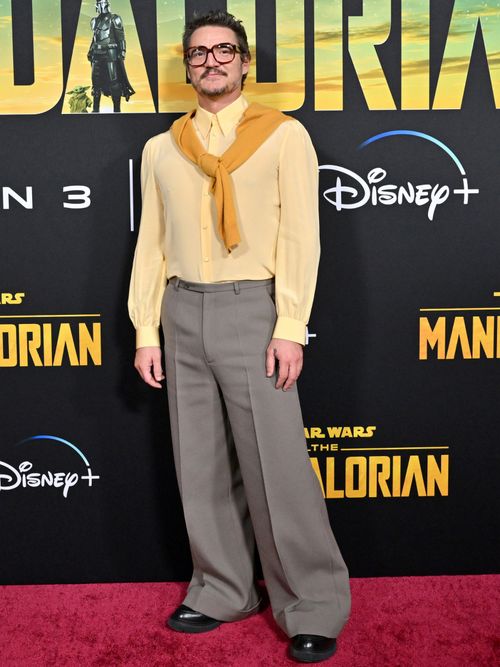 Pedro Pascal attends the Los Angeles Premiere of The Mandalorian Season 3 wearing tortoiseshell square sunglasses, a yellow jumper draped over his shoulders, a pale yellow shirt, flared grey slacks, and black derbys.