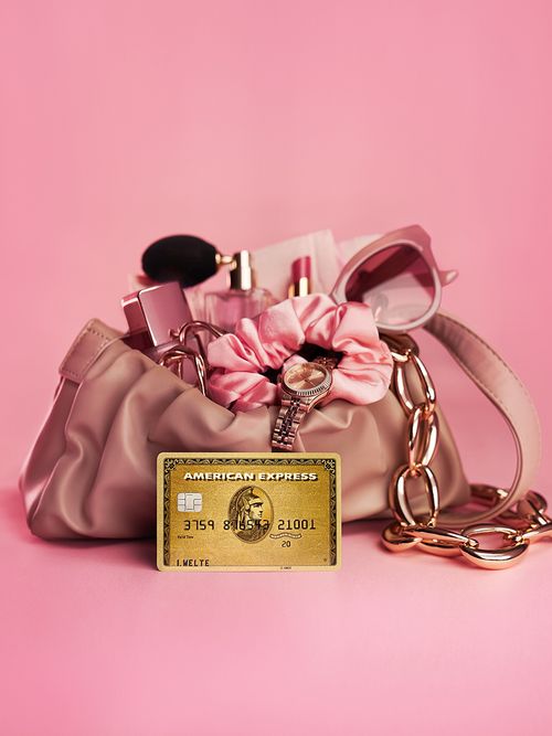 Pinke prall gefüllte Handtasche mit einem rosegoldenen Kettenriemen. Davor steht eine goldene American Express Kreditkarte.