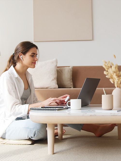 Frau sitzt im Schneidersitz am Boden vor ihrem Wohnzimmer-Tisch und arbeitet am Laptop.
