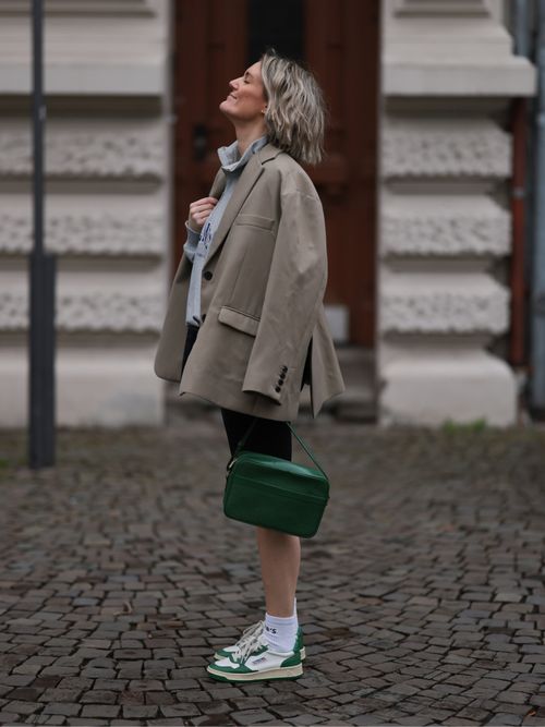 Victoria Thomas trägt einen grauen Blazer, blaue Bluse, schwarze Shorts, grüne Tasche und weiße Sneaker mit grünem Akzentstreifen in Köln.