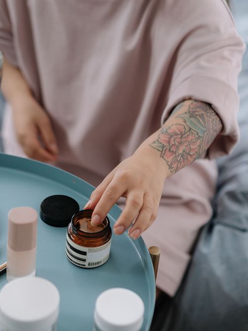 Frau in rosa T-Shirt und mit Tattoo auf dem Unterarm greift mit dem Zeigefinger in eine Cremedose.