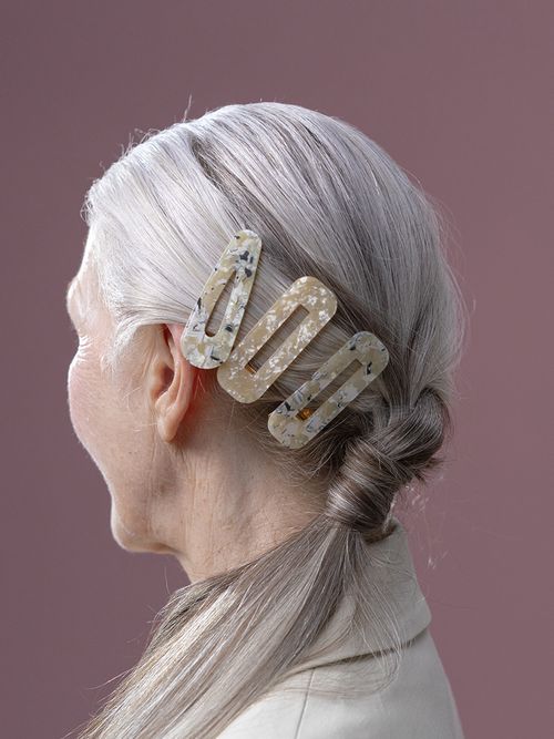 Frau mit grauen langen Haaren, die zum Pferdeschwanz gebunden sind und 3 große Haarklammern halten.