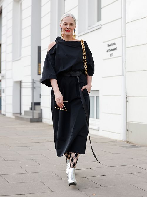 Frau mit grauen zusammengebundenen Haaren in einem schwarzen Kleid und goldenen Accessoires.