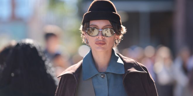 Street Style Look von der New York Fashion Week, wo ein Gast eine blaue Bluse, Cardigan, braune Jacke, Sonnenbrille und Mütze trägt.